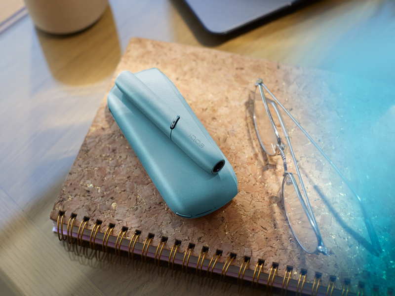Turquoise IQOS Originals Duo device on a notebook next to glasses. 
