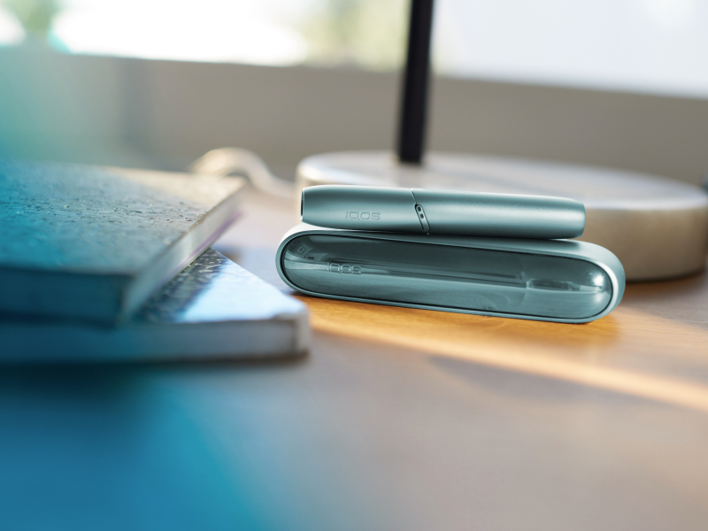 IQOS Originals Duo Holder and Pocket Charger in turquoise color on a desk, next to notebooks.