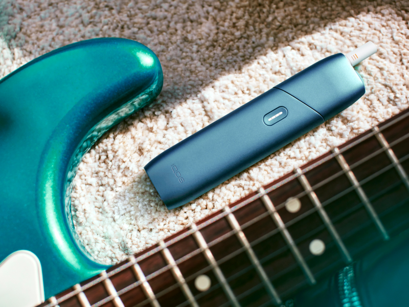"IQOS Originals One heated tobacco device, next to a turquoise guitar.