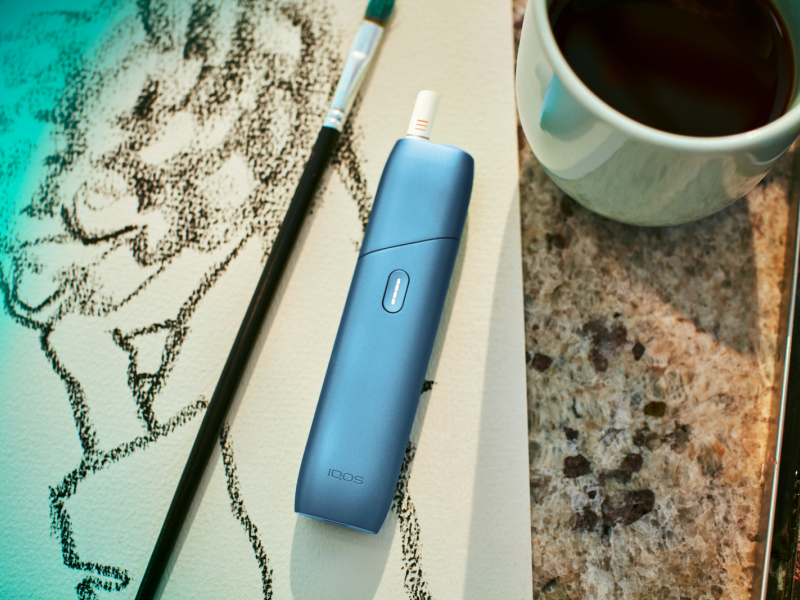 Turquoise IQOS Originals One device on a painting, next to a coffee cup.